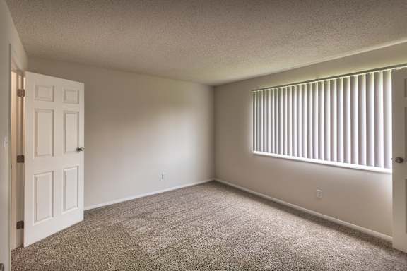 Bedroom at Village East Apartments with carpeted flooring, neutral walls, vertical blinds, and soft natural light from the window.