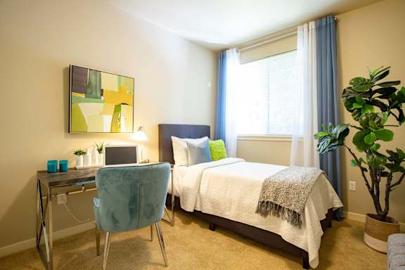 A bedroom with a bed, desk, chair, and potted plant.