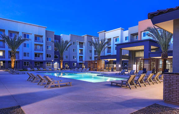 A swimming pool surrounded by lounge chairs and palm trees in front of apartment buildings.