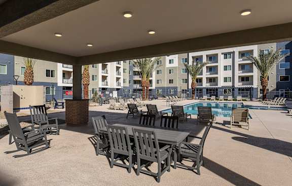 A patio with chairs and a table is in front of a pool and apartment building.