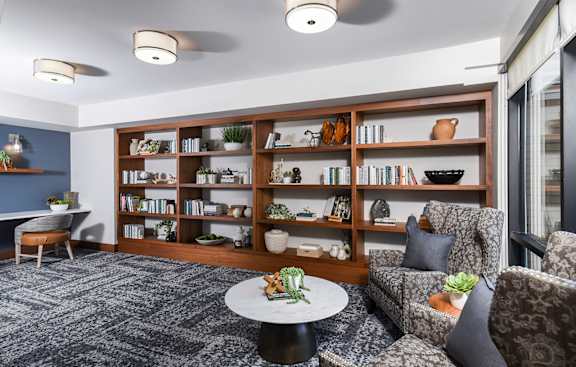 A living room with a grey carpet and a bookshelf filled with books and decorative items.