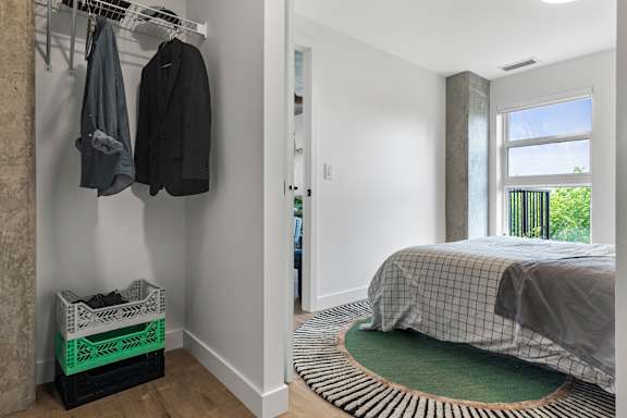 A bedroom with a bed, a coat rack with two coats hanging, and a window with a view of a balcony.