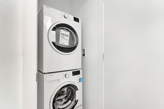 A white front load washing machine with a black door is placed in a white room.