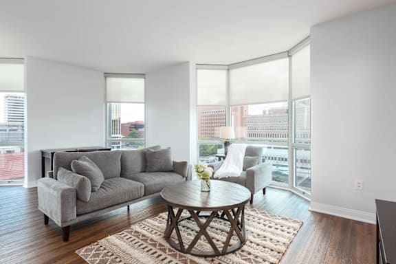 A living room with a grey couch, a coffee table and a large window at Clayton on the Park Apartments, Clayton 63105