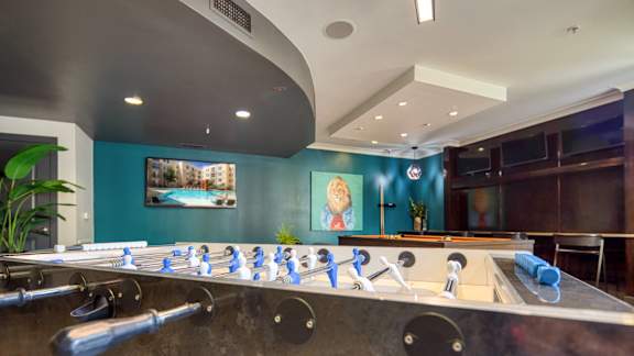 A foosball table is in the middle of a room with a blue wall and a painting hanging above it.