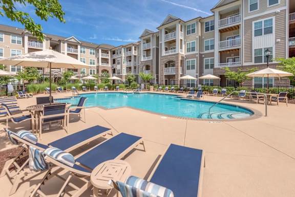 A large swimming pool surrounded by sun loungers and umbrellas in front of apartment buildings.