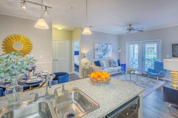 A kitchen with a sink, a counter, a fridge, a fan, a chair, a table, a vase, a bowl, and a window.