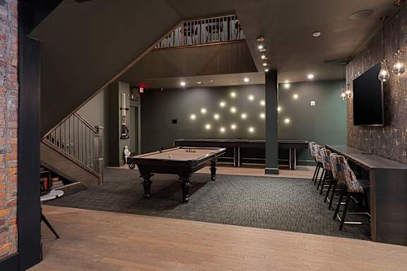 A pool table is in the middle of a room with a brick wall and a staircase.