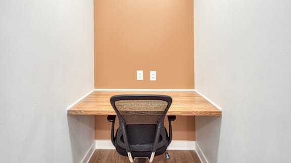A black chair is in front of a wooden desk with a white wall and two switches on the wall.
