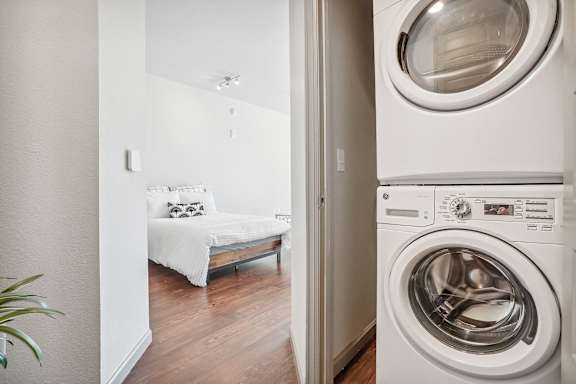 A washing machine is in the foreground of a room with a bed in the background.