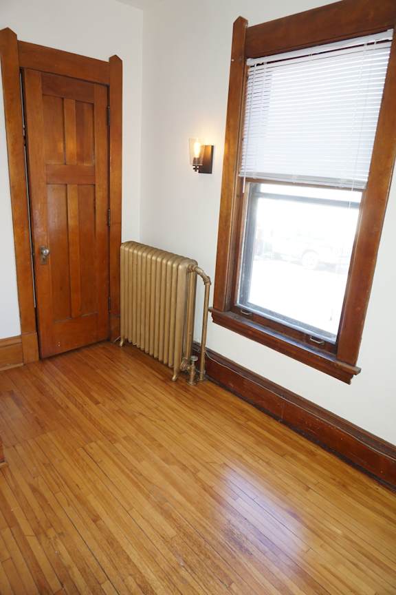A room with a wooden floor, a door, a window with blinds, and a wall-mounted light fixture.