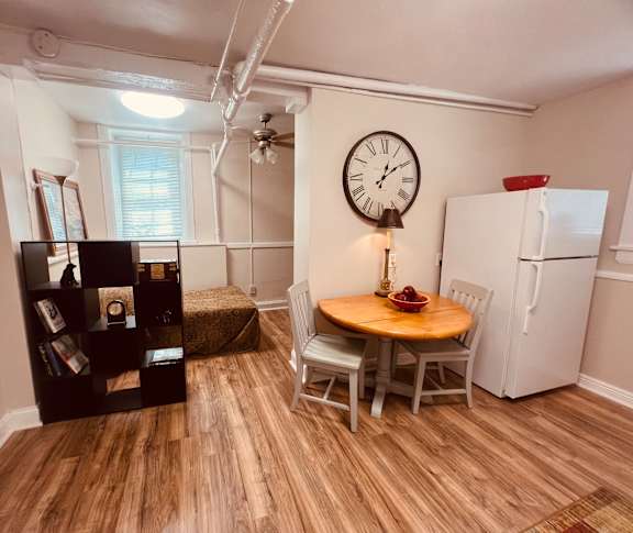 A room with a wooden table, chairs, a clock, a bookshelf, and a refrigerator.