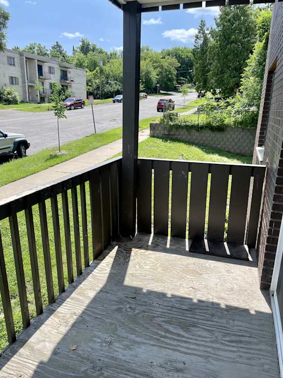 A balcony with a wooden floor and a black railing overlooks a street with cars and buildings.
