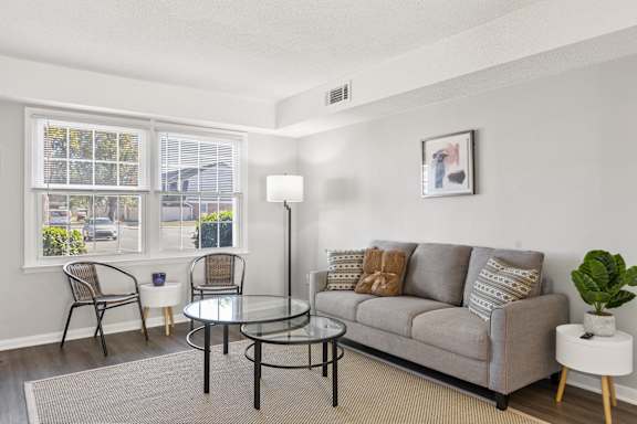 A living room with a grey couch, a glass table, and a window with a view of the street.