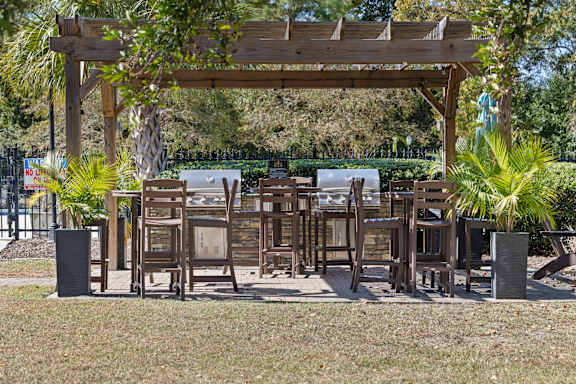 A wooden pergola with chairs and tables is surrounded by greenery.