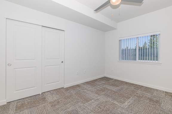 A room with a carpeted floor, a ceiling fan, and a window with blinds.