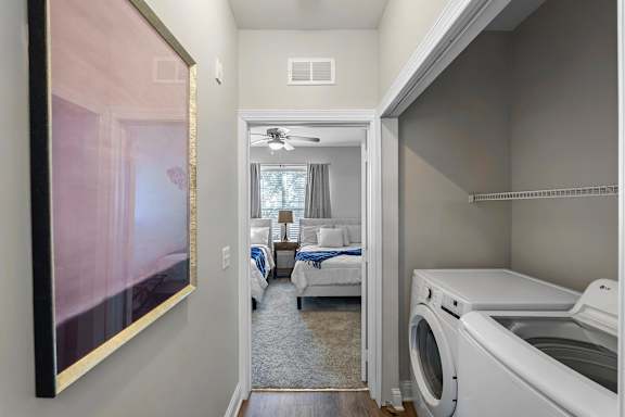 A white washing machine is in a laundry room with a bedroom in the background.