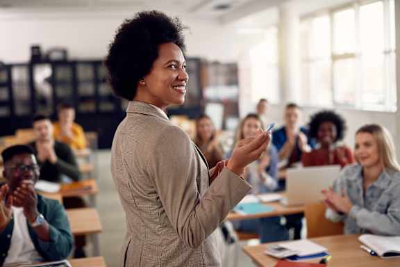A woman in a beige blazer is standing in front of a classroom full of students who are clapping.