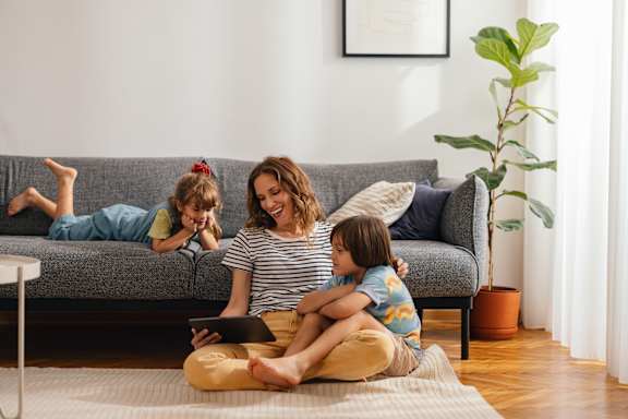 A woman and two children are sitting on the floor in front of a grey couch.