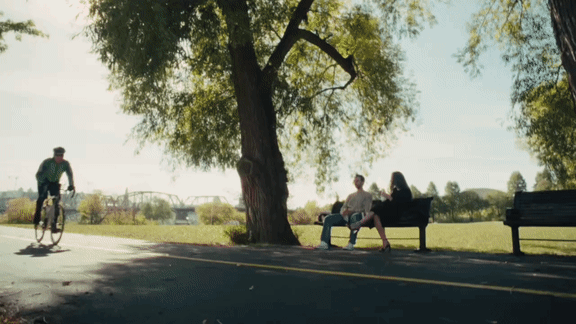 A man is riding a bicycle on the road while two people are sitting on a bench.