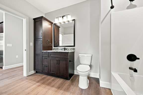 A white toilet sits in a bathroom with a wooden floor and a white tub.