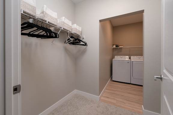 A white closet with clothes hanging and a laundry room in the background.