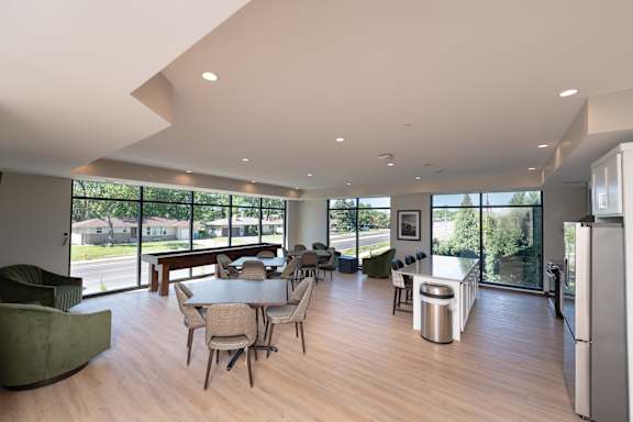 A modern community room kitchen with a dining table, comfortable chairs and shuffle board game.