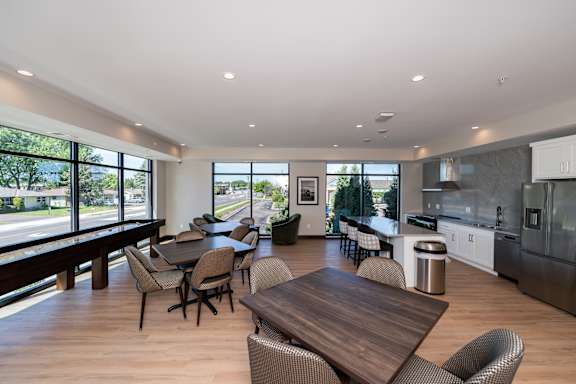 A modern community room kitchen with a dining table, comfortable chairs and shuffle board game.