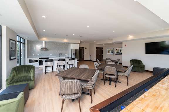 A modern community room kitchen with a dining table, comfortable chairs and shuffle board game.