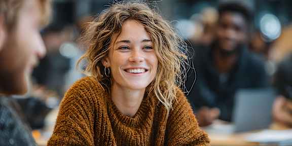 A woman with curly hair is wearing a brown sweater and smiling.