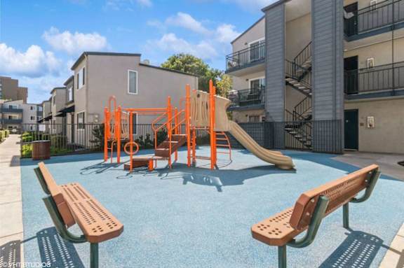 A playground with a slide, swings, and benches in front of a building.