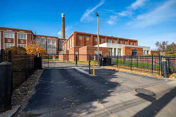 A brick building with a tall chimney is behind a black fence.