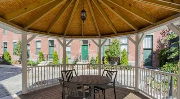 A wooden gazebo with a table and chairs is in the foreground with a building in the background.