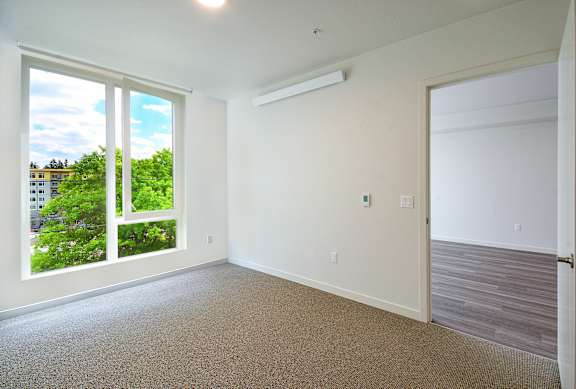 A room with a carpet floor, a window with a view of trees and buildings outside, and a closed door.