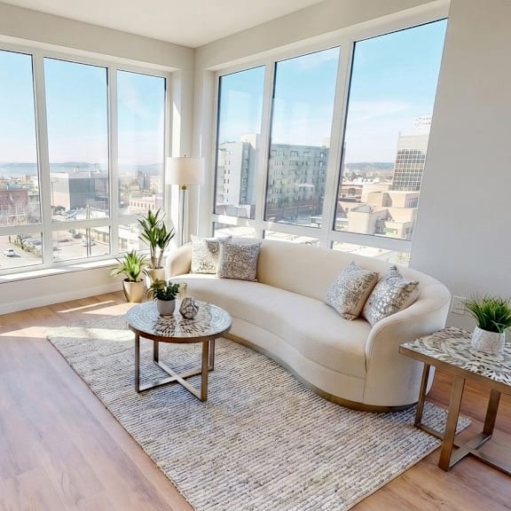 A living room with a white couch, a coffee table, and a large window overlooking a cityscape.