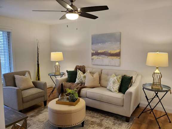 A living room with a beige couch, a chair, a table with a lamp, and a painting on the wall.