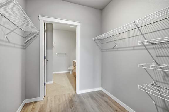 A white closet with shelves and an open door leading to a bathroom.