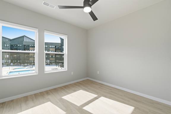A room with a ceiling fan and two windows overlooking a pool and buildings.