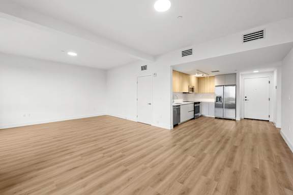 A room with wooden flooring and a kitchen area in the background.