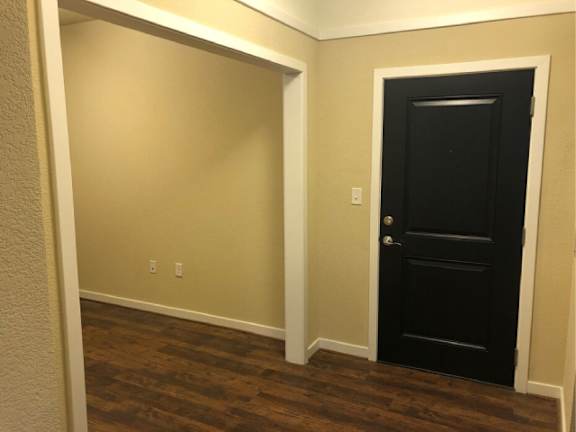 A black door is on the right side of a hallway with wood floors and beige walls.
