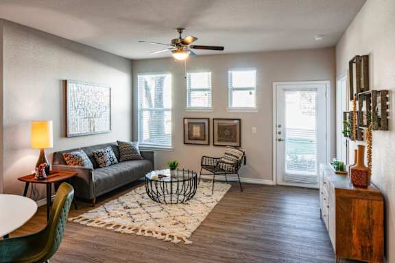 A living room with a grey couch, a green chair, a white table, a rug, a ceiling fan, and a door leading to a balcony.