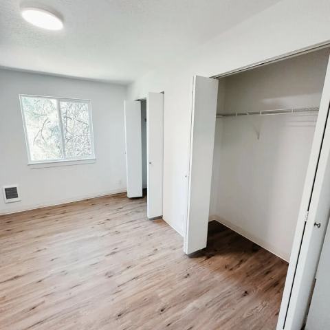 A room with white walls and wooden flooring, featuring a large open closet and a window.