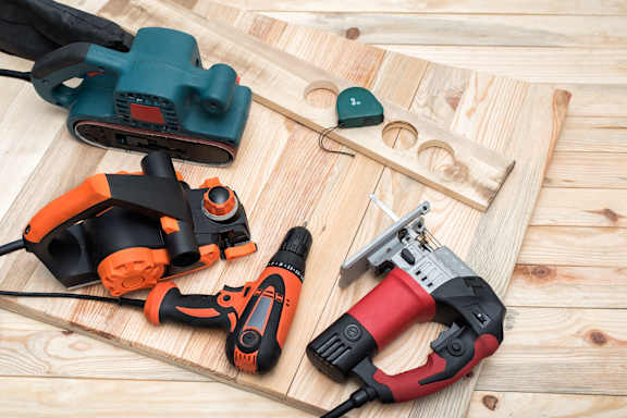 A collection of power tools including a jigsaw, drill, and nail gun are laid out on a wooden surface.