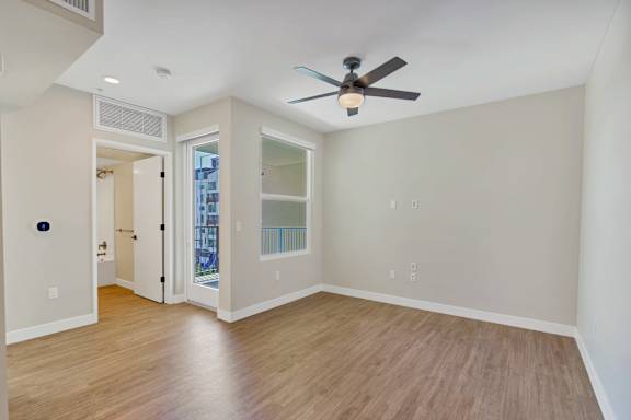 A room with a ceiling fan and light, hardwood floors, and a door leading to a balcony.