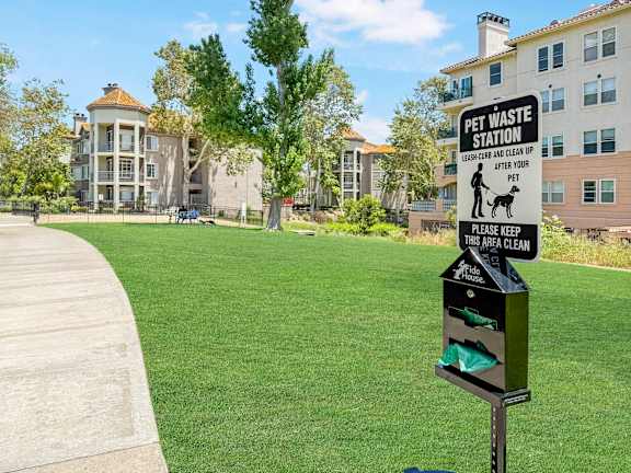A sign for a pet waste station is on a pole in a grassy area.
