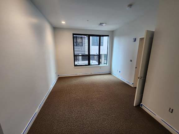 A room with brown carpeting and a window overlooking a building.