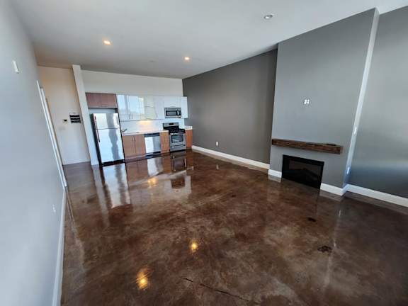 A large, empty room with a fireplace and a kitchen area in the background.