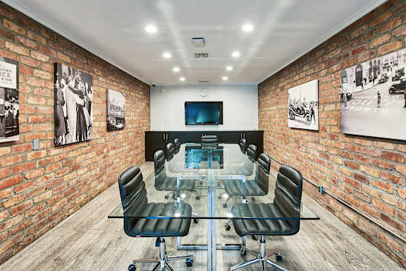 A conference room with a glass table and chairs, a television, and framed pictures on the brick walls at The Meadows Apartments, Culver City , CA