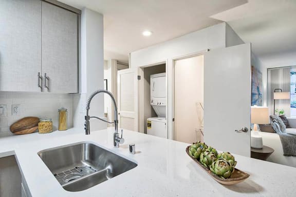 A modern kitchen with a stainless steel sink and a wooden tray of artichokes on the counter at The Meadows Apartments, Culver City