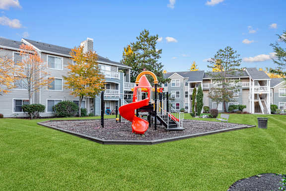 A playground with a slide in the middle of a grassy area in front of apartment buildings.